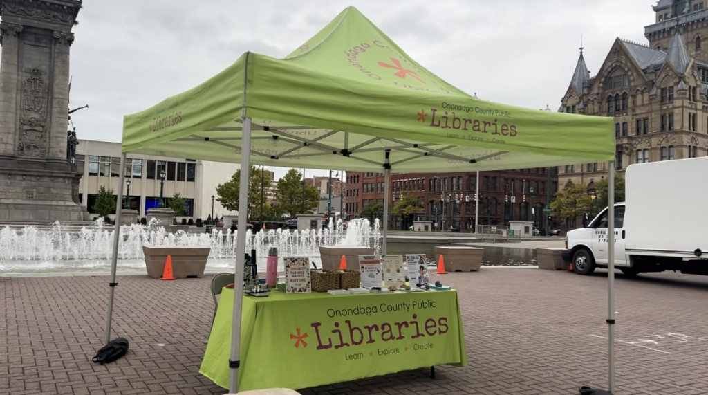 A booth representing the Onondaga Public Libraries in front of a fountain in Downtown Syracuse.