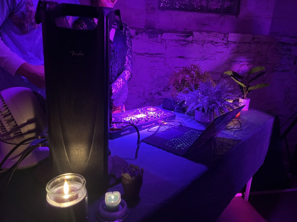 A DJ mix board with a laptop next to candles and plants under colorful lights.