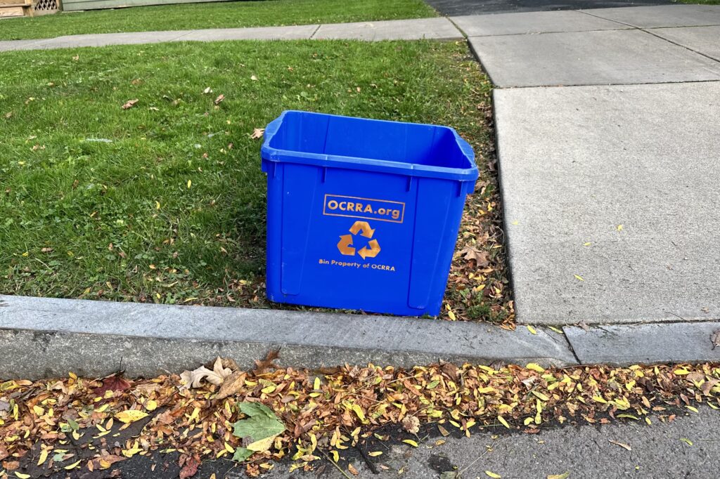 Small blue recycling bin sits on a front yard.