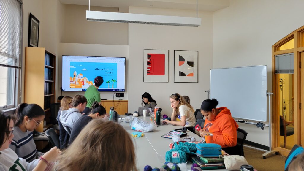 Students sitting around a table crocheting and listening to a classmate present at the front of the room about a crochet charity organization.