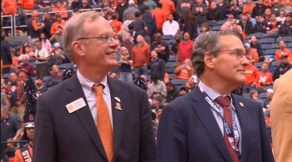 Syracuse University Chancellor Kent Syverud, wearing a navy blue suit with an orange tie, stands next to athletic director John Wildhack.