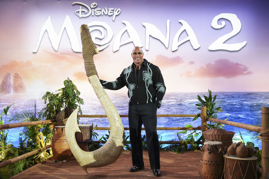 Dwayne Johnson stands next to a large fish hook in front of an ocean background. 