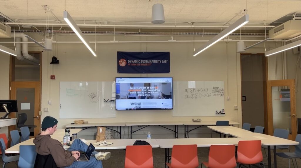 A classroom space for lab students to work with a student at the round table with orange chairs, TV screen and whiteboard.