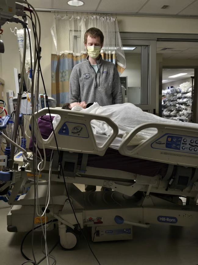 Jamie lies in a hospital bed with his eyes closed as a doctor looks over him in Rochester, N.Y. 