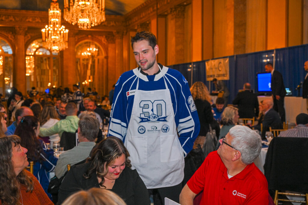 A hockey player wearing a blue Syracuse Crunch jersey interacts with people at a table.