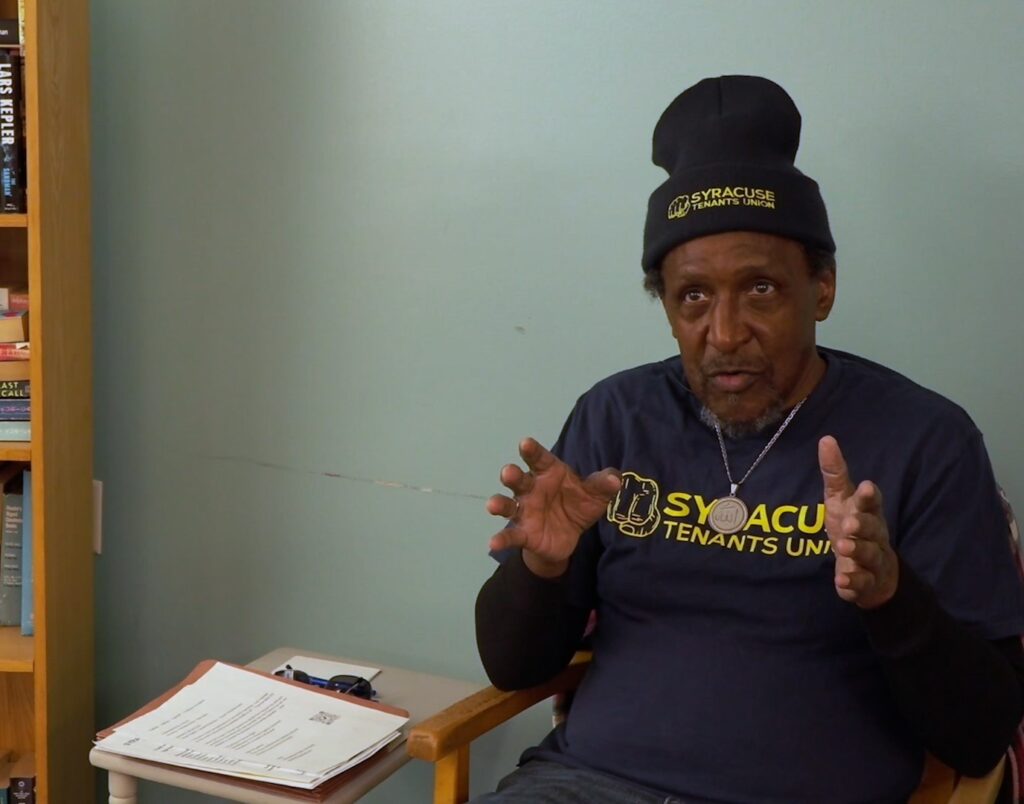 A black man in his late 60's in the middle of an interview. He's wearing a Syracuse Tenants Union hat and shirt. 