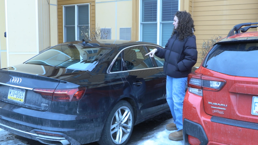 Olivia Meier points at back window of a black Audi car in a parking lot.