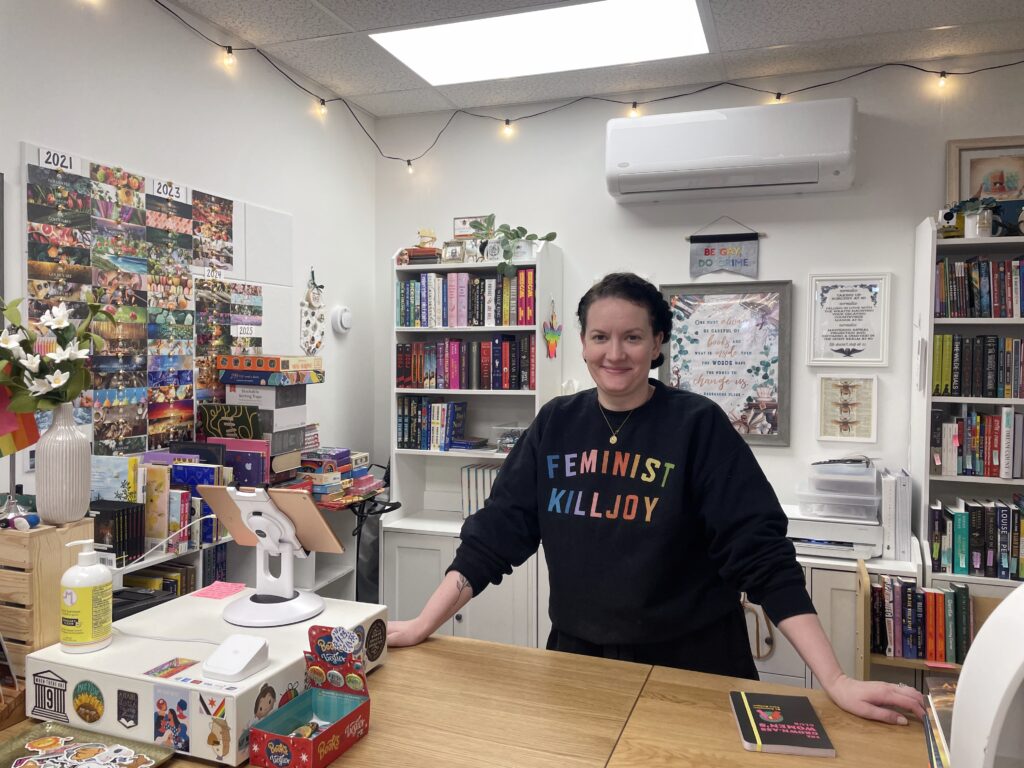 Women with black sweater leaning on wooden desk with bookshelves behind her.