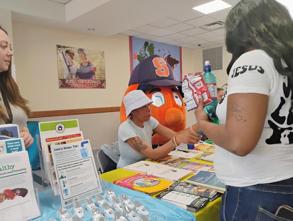 Volunteers share lead safety information and distribute water testing kits at a colorful resource table. Syracuse University’s mascot, Otto the Orange, stands behind the table, adding energy and school spirit as attendees engage with the materials.