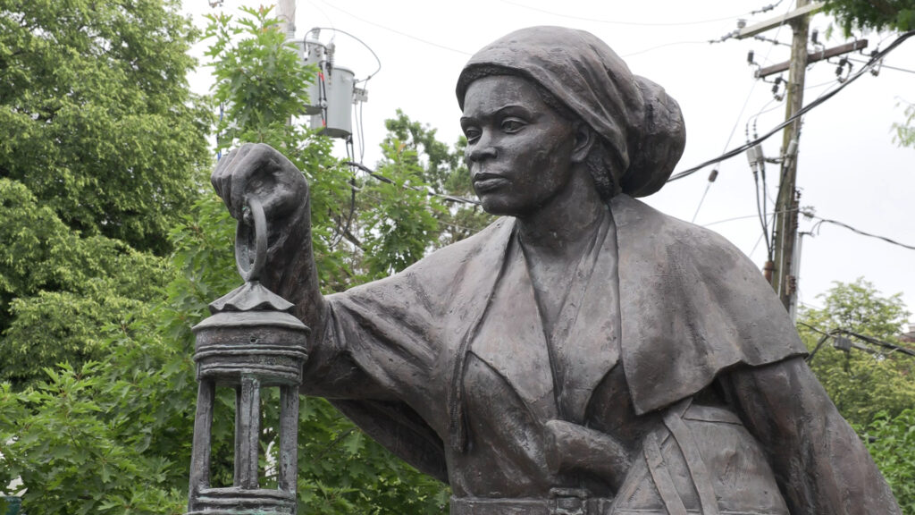 Harriet Tubman statue holding a lantern in the plaza of the NYS Equal Rights Heritage Center with trees and light poles in the background. 