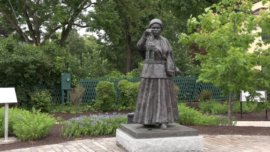 Harriet Tubman statue holding a lantern in the plaza of the NYS Equal Rights Heritage Center with trees and light poles in the background. 