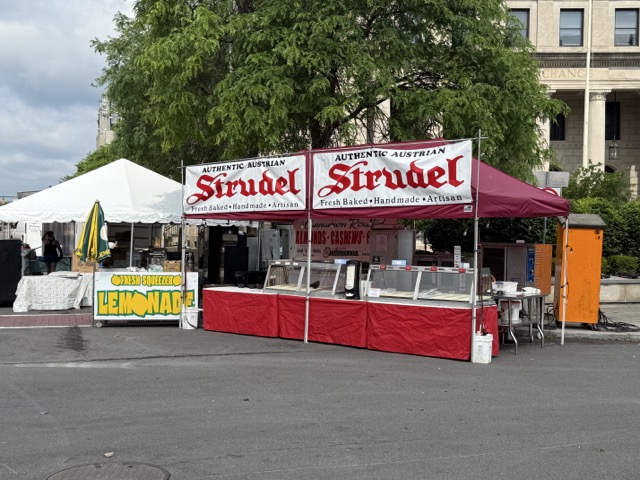 A vendor booth with red and white signage reading “Authentic Austrian Strudel” is set up at the Syracuse Polish Festival, alongside a yellow “Fresh Squeezed Lemonade” stand.