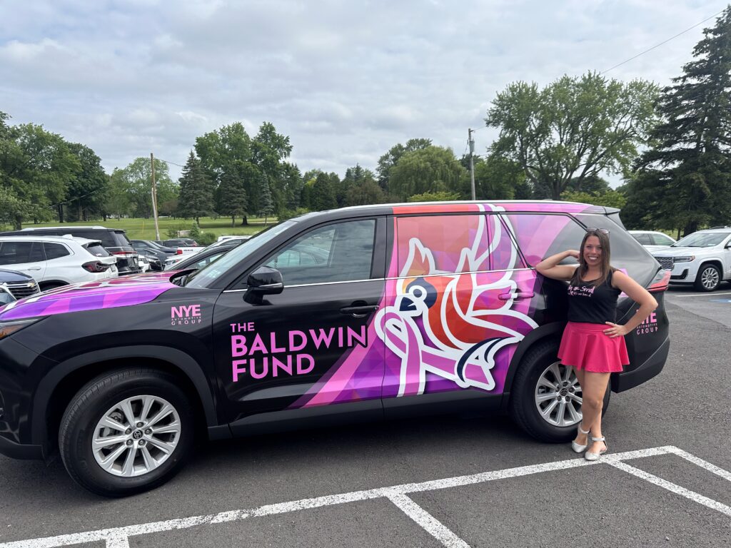 Breast cancer survivor Courtnie Walker pictured next to The Baldwin's Fund vehicle. The vehicle is black with and exterior design of a bird and pink ribbon. The colors are pink, orange and yellow.