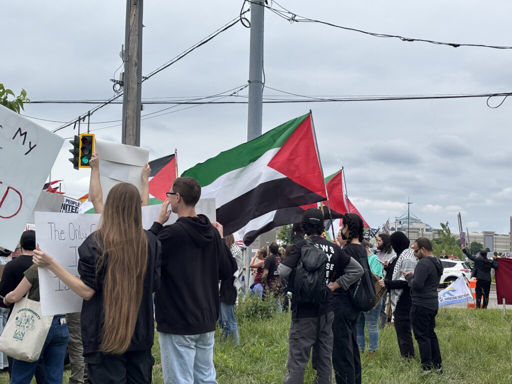 A crowd of protesters gathers under traffic lights, holding Palestinian, pride, and various activist flags.