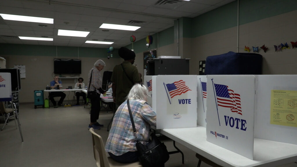 A woman sits at a voting booth to fill out her ballot. There are several blockers up to provide privacy for the people voting. They say vote on the side with an American flag.