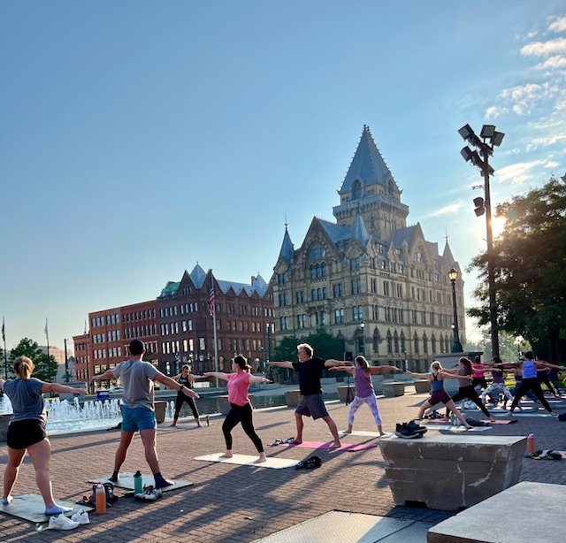 People doing outdoor yoga in a city square with a historic building and fountain in the background. © 2025 Maggie Robinson