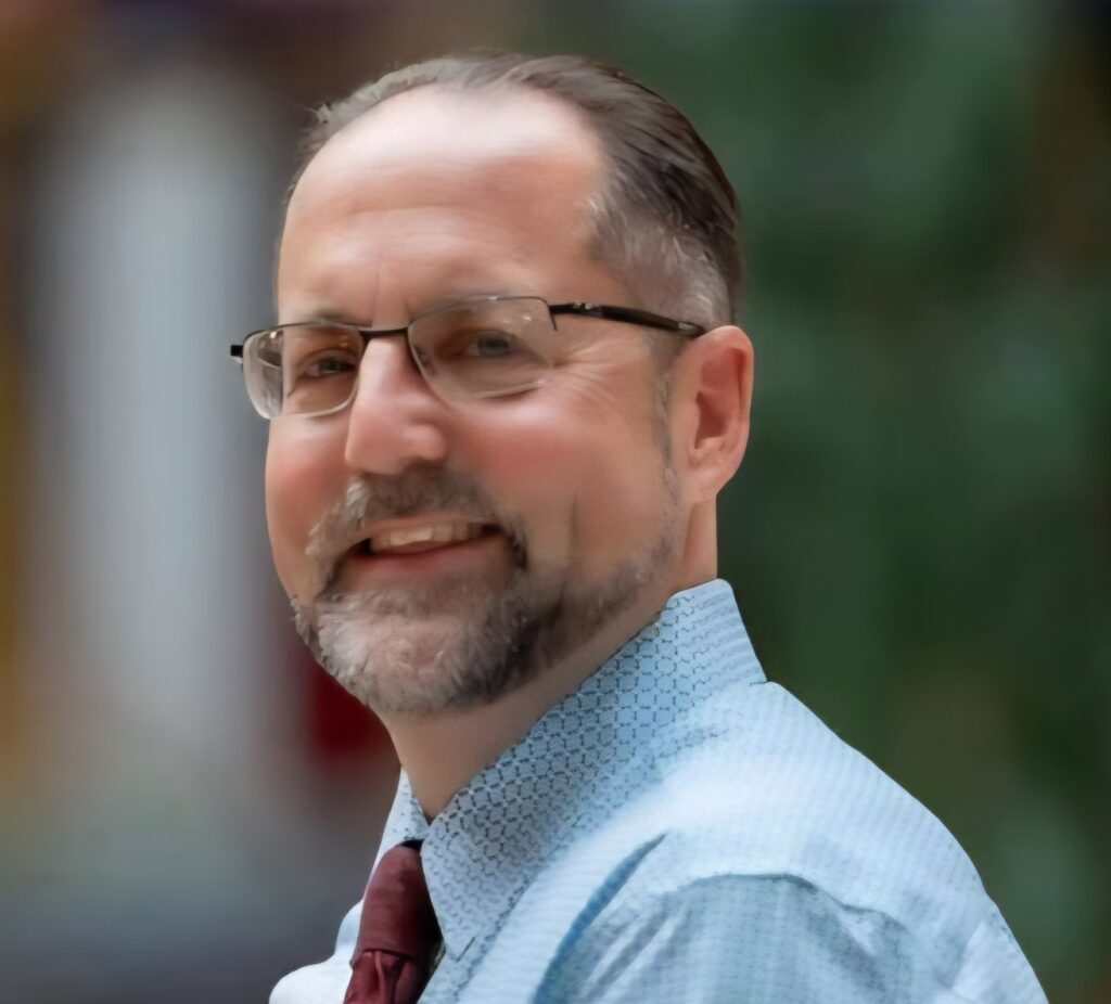 A man wearing glasses, a blue collared shirt and red tie smiles at the camera.