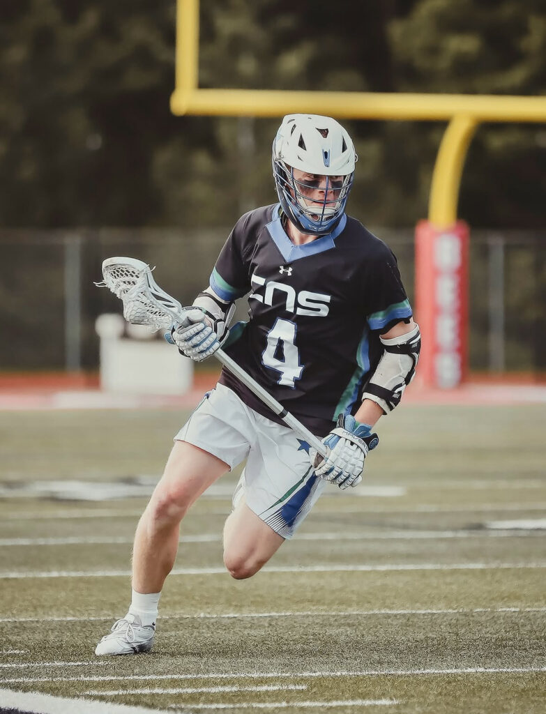 Lacrosse player wearing a black jersey, a white helmet with a star and gray pants carries a lacrosse ball in his stick on a high school field.