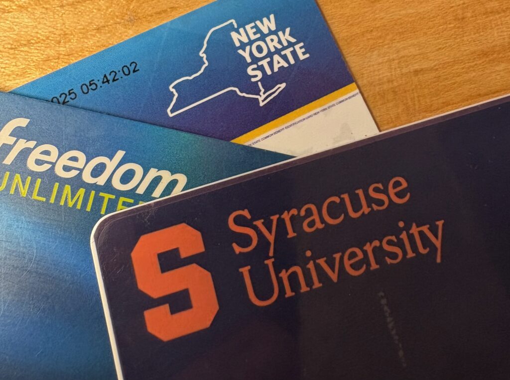 Three cards piled on top of one another on a wooden surface. The bottom is a New York State EBT card, the middle is a Freedom Unlimited credit card, and the top is a Syracuse University ID card.