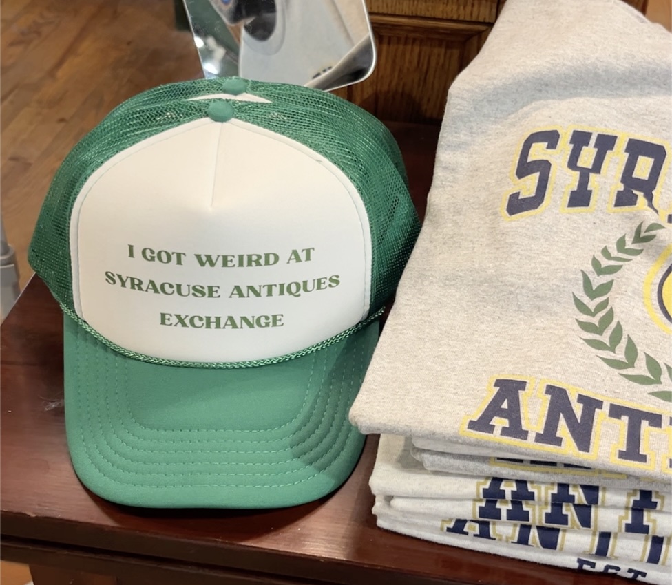 Syracuse Antiques Exchange original merchandise. A green trucker hat with green text that reads "I got weird at Syracuse Antiques Exhange" and a grey crew neck with varisty-style letter text that reads "syracuse antiques" in navy and yellow.