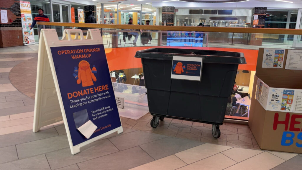 A gray, Operation Orange Warmup collection bin and sign sitting in the middle of the Schine Student Center.