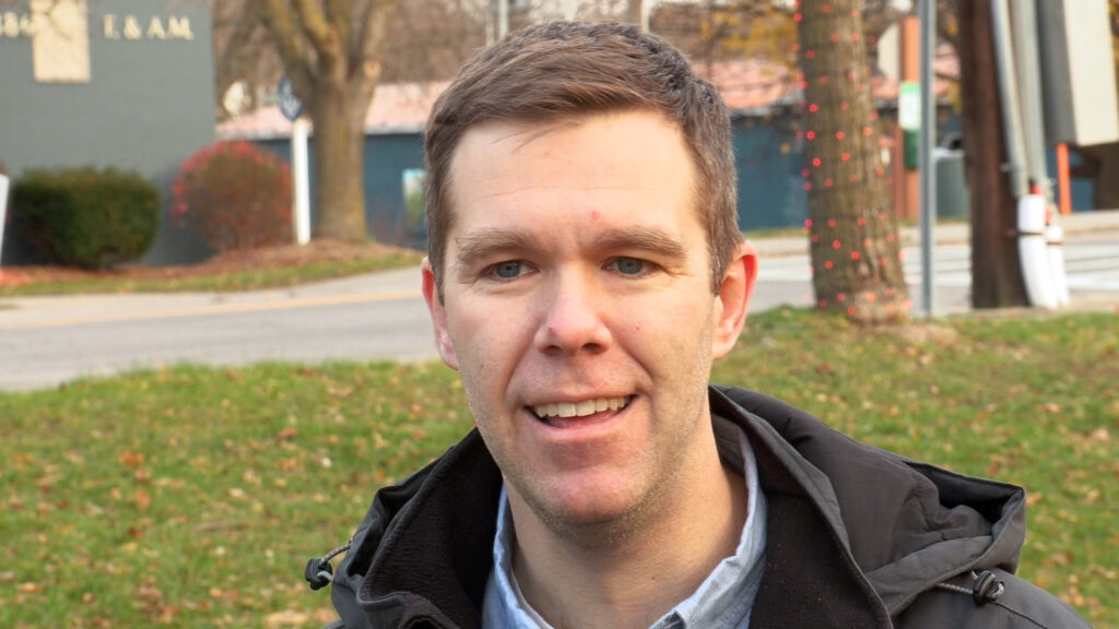 Ken Bush III stands on grass in front of the blue Jordan Masonic Lodge which is the starting point of the race.