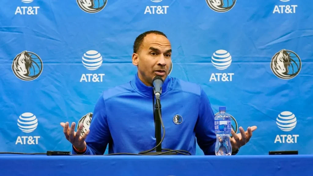 In front of a blue AT&T backgorund, Nico Harrison sits in a blue shirt at a press conference speaking into a microphone.