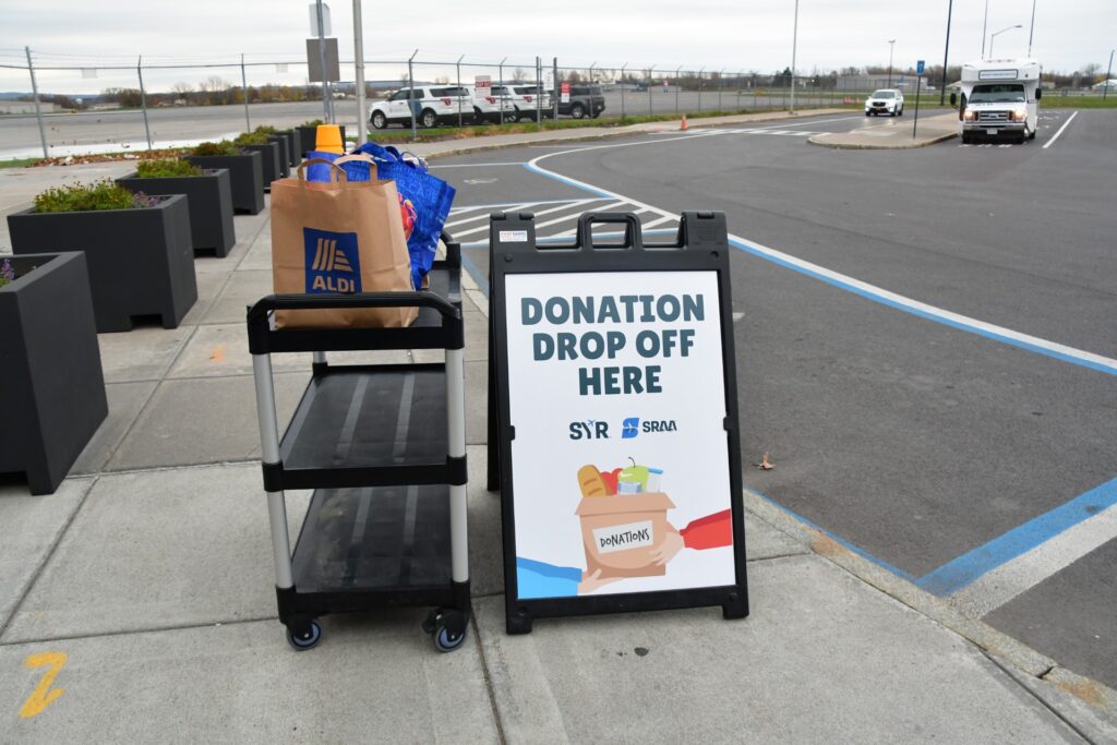A sign near a curb of a pick up area reading "Donation Drop Off Here" next to a cart with several bags of food.