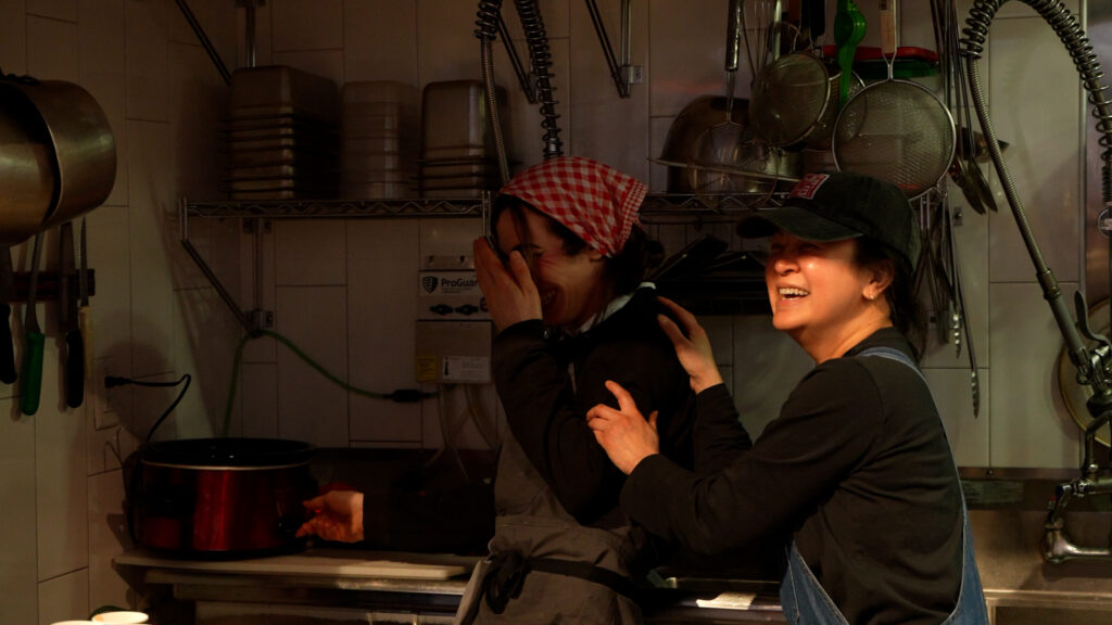 Two women laugh while in a kitchen. The woman on the right is playfully grabbing the arm of the woman on the left