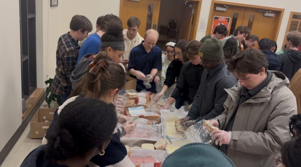 Students gather around a table to form an assembly line, making bologna and cheese sandwiches.