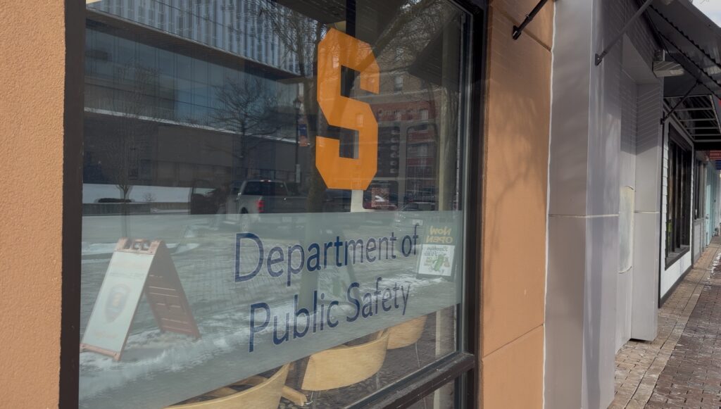 A window of Syracuse University’s Department of Public Safety office, displaying a large orange “S” and the department’s name, with reflections of the street and buildings visible on the glass.