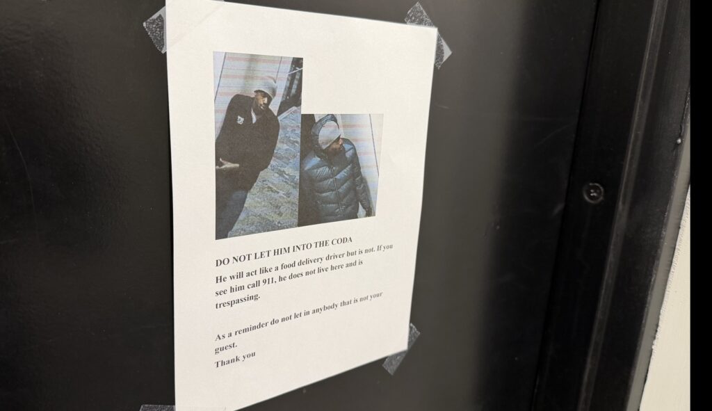 A printed warning flyer taped to a black door shows two security camera photos of a man wearing a winter hat and dark jacket. The flyer reads “DO NOT LET HIM INTO THE CODA” and warns that he has pretended to be a food delivery driver to enter the building.