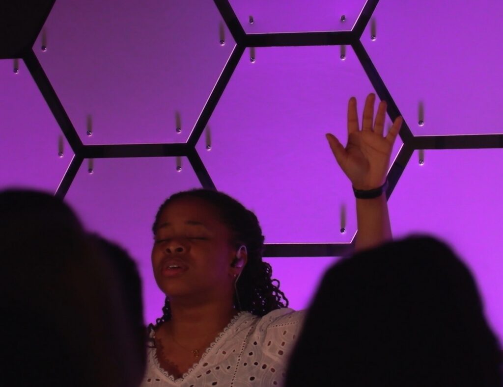 A young woman is singing at The Salt Company worship with her hand in the air and eyes closed.