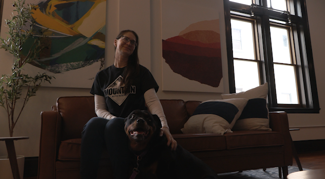 A rescue dog owner sits on a couhc next to her dog ans smiles at a point in the distance.
