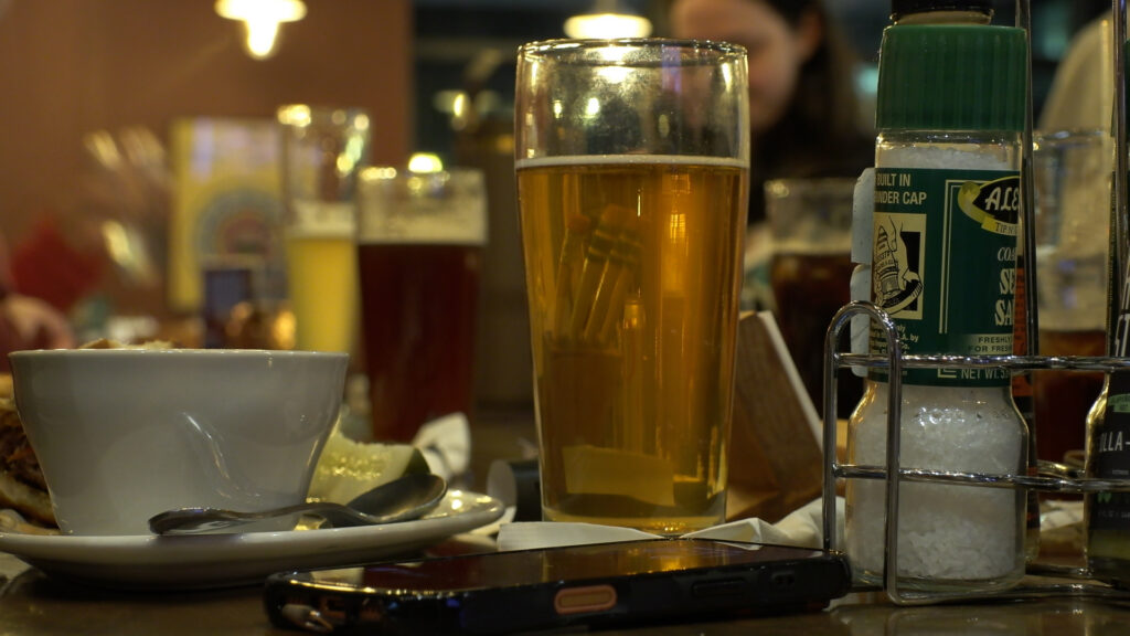 A beer on a table surrounded by food and other beverages.