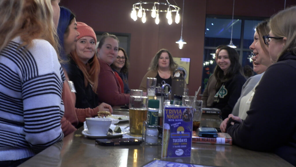 Group of women around a table with beers, books, and food. 