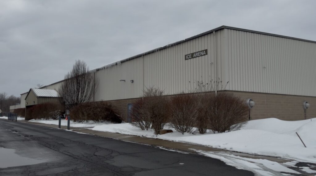 A snowy exterior view of the Three Rivers Athletic Complex, showing the metal-sided building with a sign that reads “Ice Arena” above leafless shrubs.