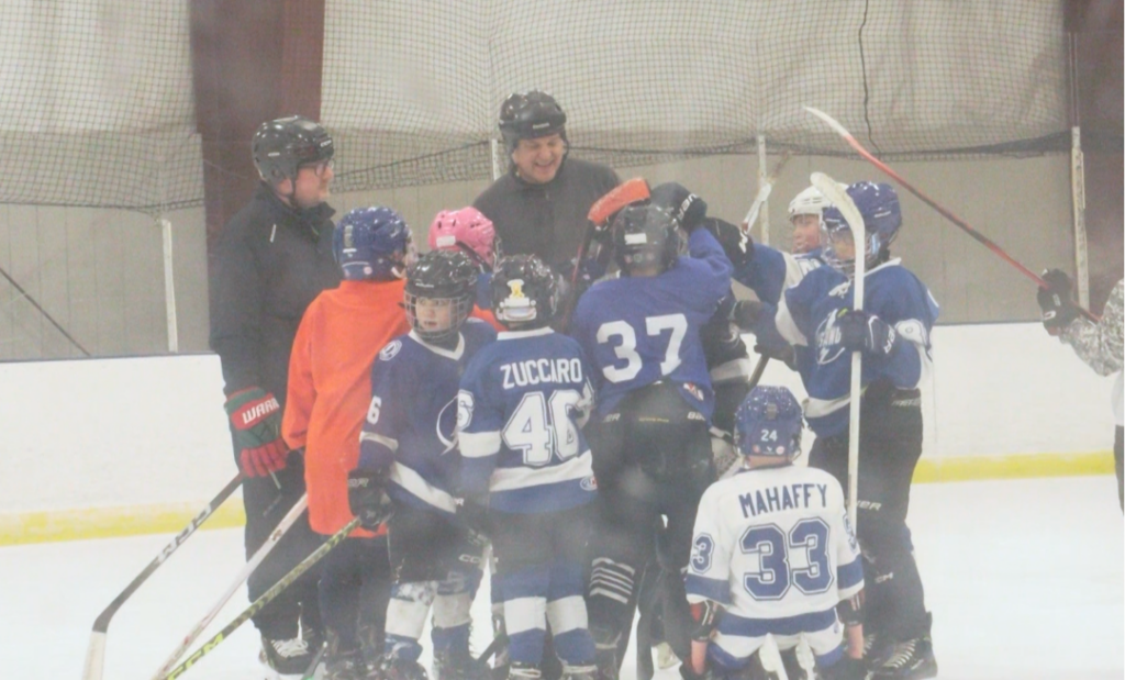 A youth hockey coach stands at center ice surrounded by a group of young players in blue and white jerseys, all gathered in a tight huddle during practice at the Three Rivers Athletic Complex.