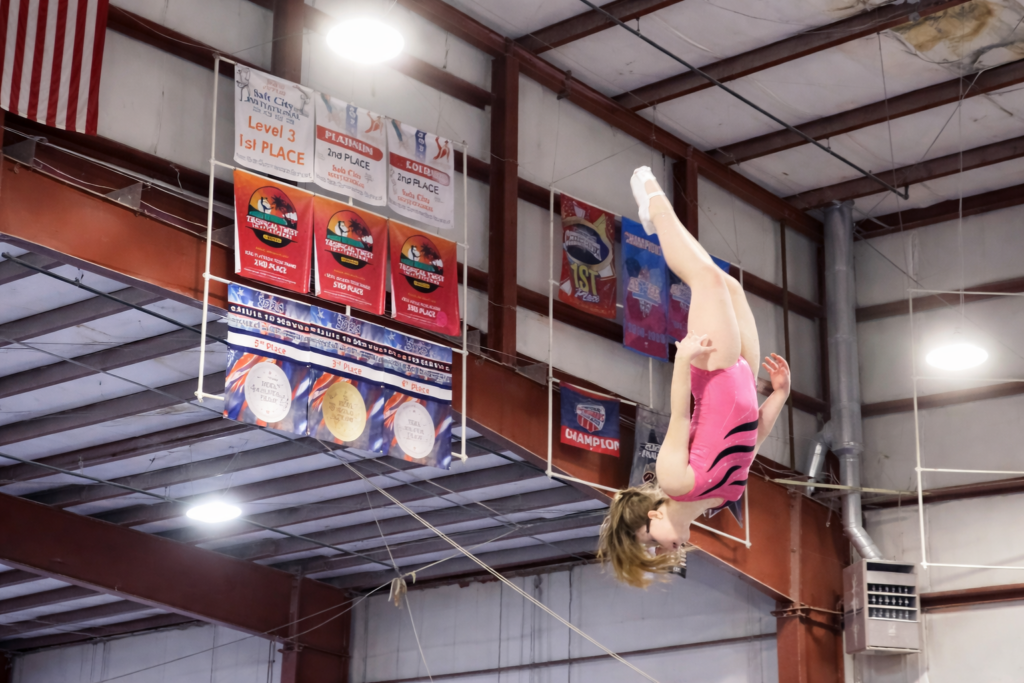 A gymnast in a pink leotard flips high in the air inside a trampoline gym with banners hanging in the background.