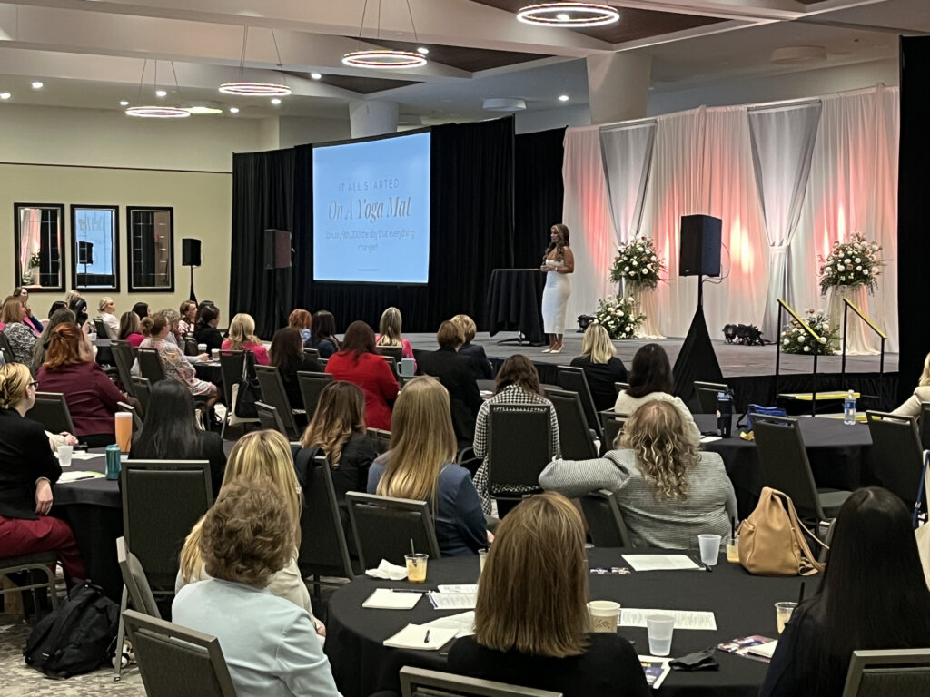 Woman on right side in white dress speaks to attendees as their keynote speaker.