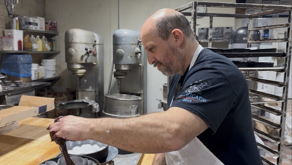 Peter Papas puts chocolate cake batter into cake pans in the bakery, with large stand mixers in the background
