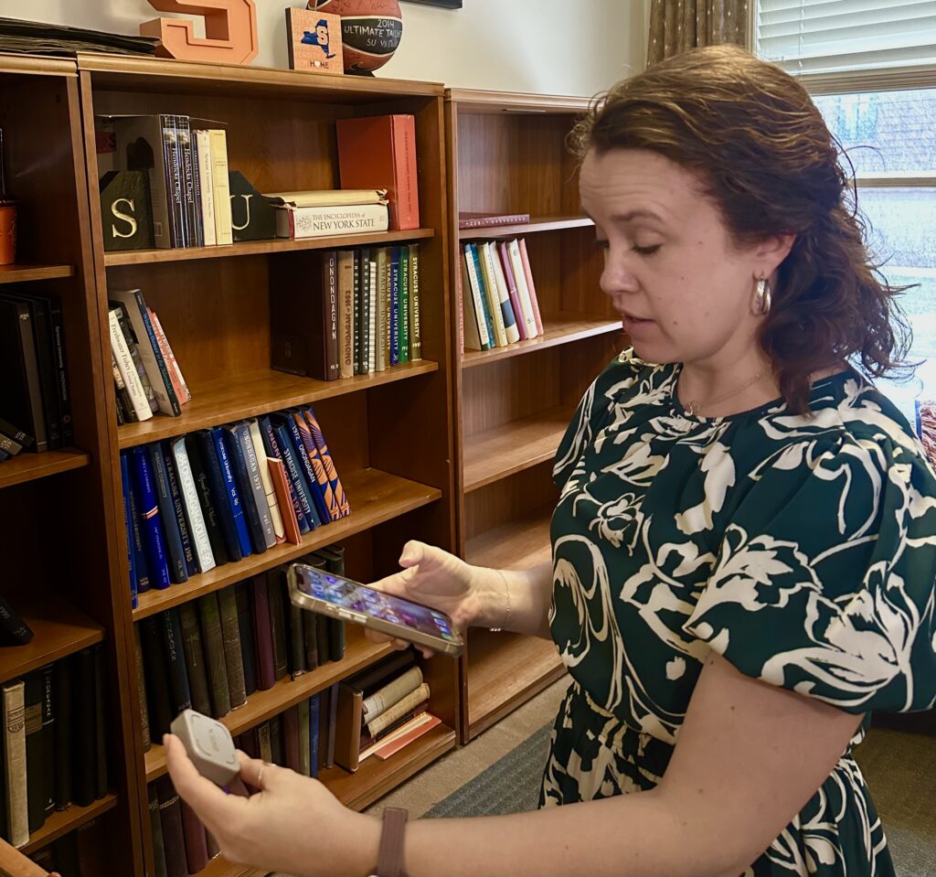 A women is holding a gray box that says "Brick" with an iPhone. There is a bookshelf behind her.