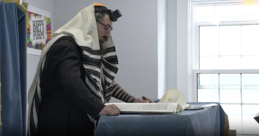 Rabbi stands in front of podium reading scroll to congregation at Syracuse Chabad. 
