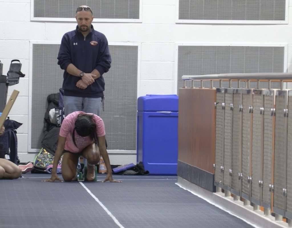A Liverpool High School sprinter prepares in starting blocks during indoor practice while coach Greg Hamilton watches from the sideline