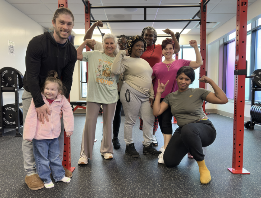 A group of seven adults and one young child pose and flex their muscles in front of a red squat rack inside a gym. Weight plates line the sides of the frame and large windows let in natural light in the background.
