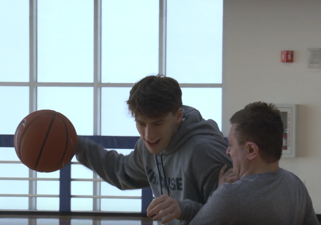 Basketball player in a Syracuse hoodie dribbles while backing down another player. Both athletes are grinning.