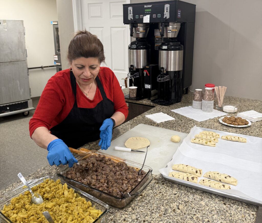 A woman wearing gloves prepares food on a kitchen counter with trays of dough and filling nearby.