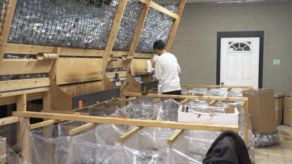 Man in white shirt sorts plastic water bottles into clear bags. He stands in fromt of a wooden structure holding all bottles.