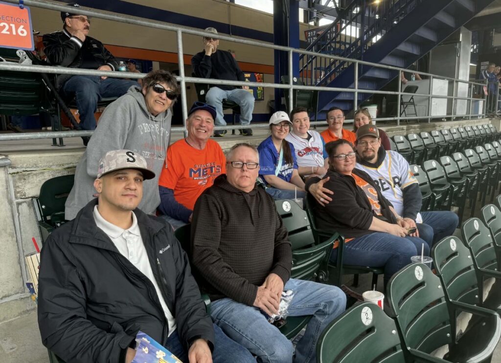 A group of family and friends sit together in two rows of seats at a baseball game, with an empty seat in the lower middle row. 