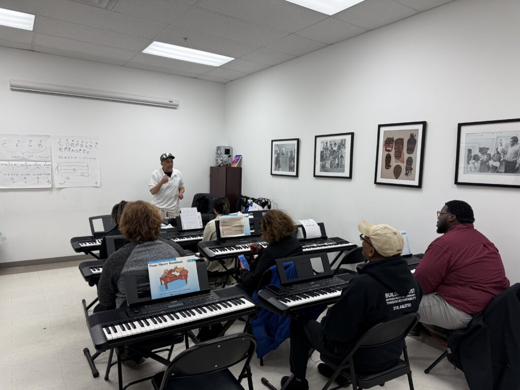 Adult piano students sit at keyboards during a beginner class at the Community Folk Art Center while instructor Bernard Wiliford teaches from the front of the room. Music theory notes are written on a whiteboard as students follow along.